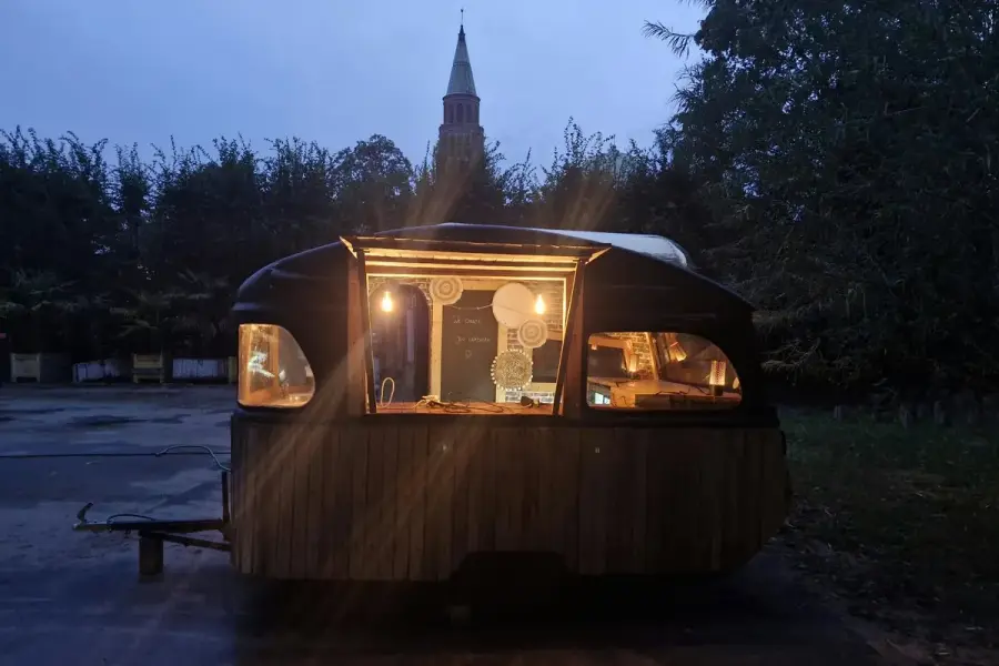 De Bobonne: a vintage wooden caravan at dusk, warmly lit, with a chalkboard inside and a tower in the background