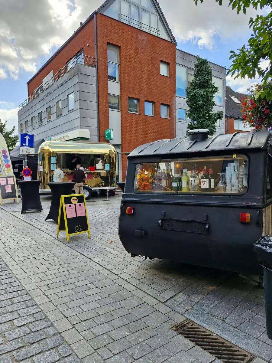 A golden Airstream and a black wooden caravan bar side by side on a street square with standing tables