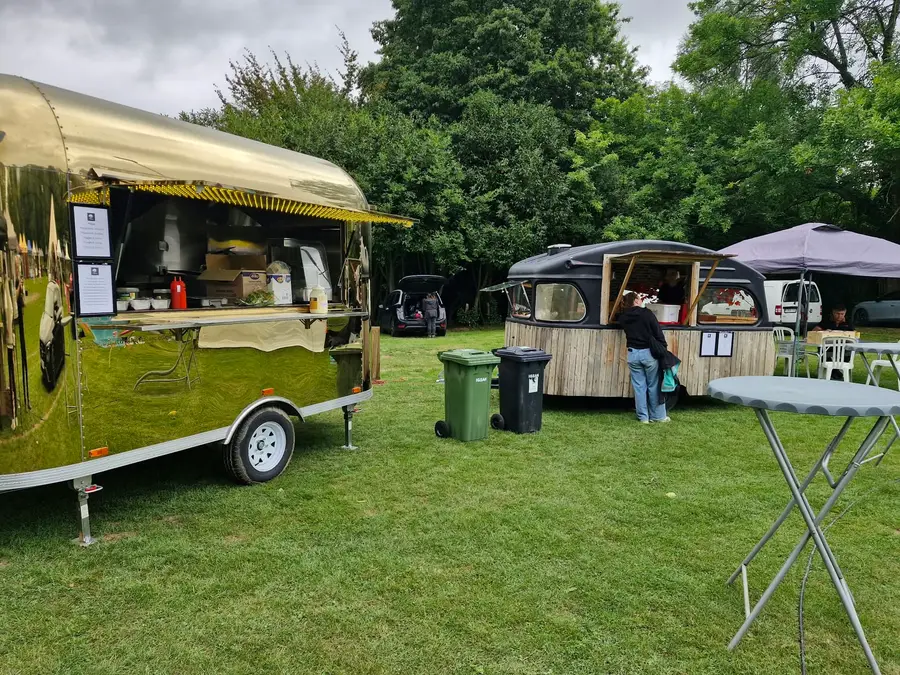 Two food trucks, a golden Airstream and a wooden caravan, on a lawn among the trees