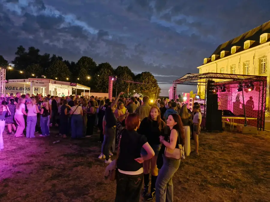 Busy evening party in a park in front of a castle, with a stage and colourful lighting